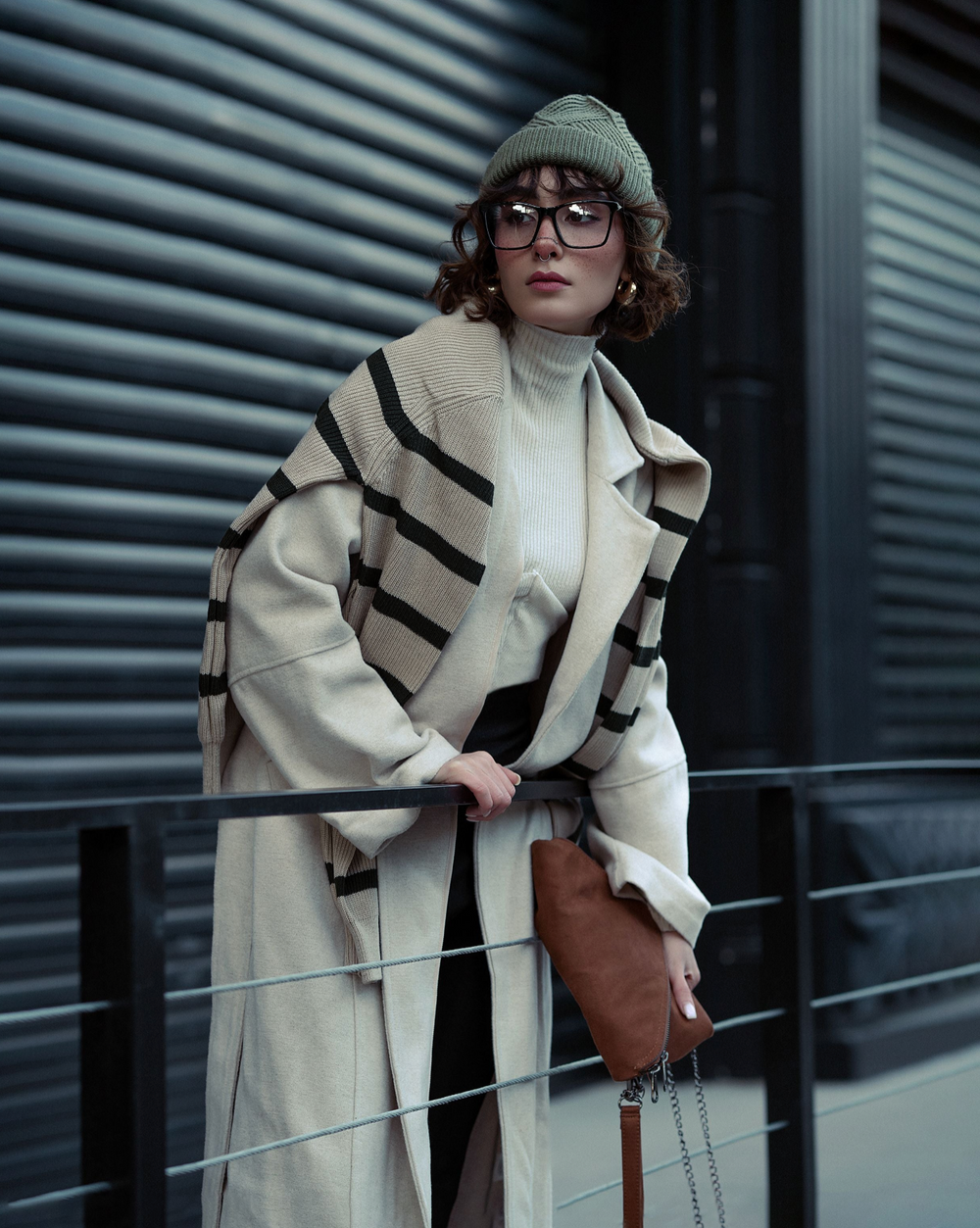 A woman wearing a beige and black striped wool coat over a cream turtleneck, paired with a green knit beanie, black-framed glasses, and a brown leather clutch, posing in front of a dark corrugated metal wall.